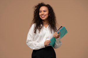 A imagem mostra uma jovem mulher sorridente com cabelo cacheado e volumoso, vestindo uma camisa branca de mangas longas e calça preta. Ela segura uma pasta de arquivos azul-clara e alguns cadernos coloridos. O fundo é de cor bege uniforme, destacando a figura da mulher. Ela parece estar em um ambiente profissional ou educacional, sugerindo uma atmosfera de confiança e prontidão. A imagem é a capa escolhida para ilustrar a matéria sobre "Como a graduação impacta a sua carreira profissional?"