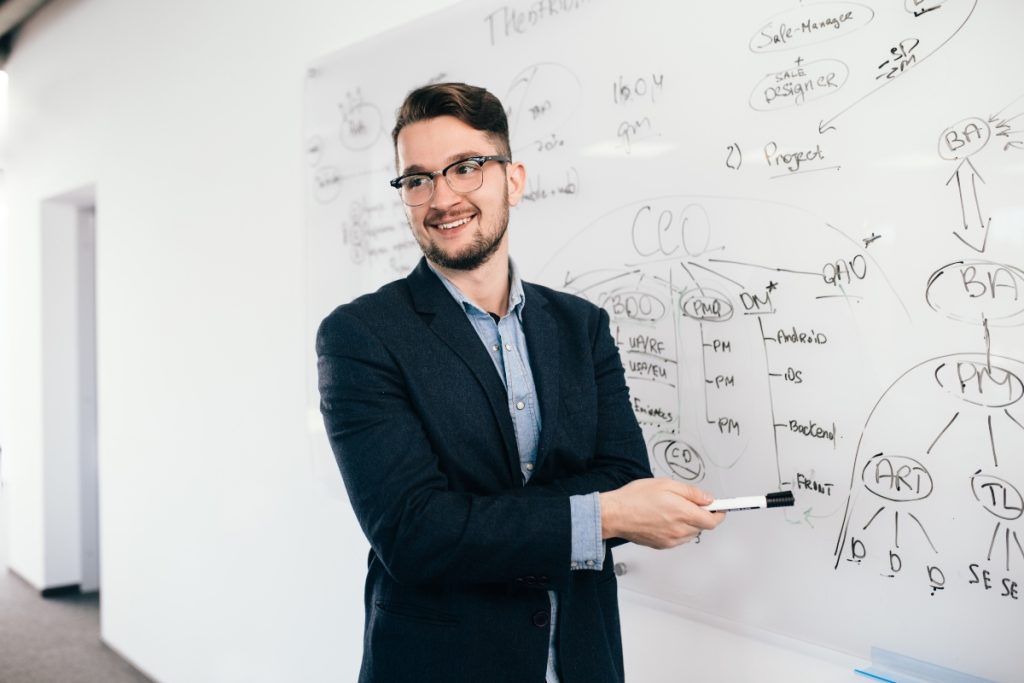 A imagem mostra um homem jovem, usando óculos e um blazer escuro sobre uma camisa jeans, sorrindo enquanto segura um marcador de quadro branco. Ele está de pé ao lado de um quadro branco coberto de diagramas e anotações. O ambiente parece ser um escritório ou sala de aula moderna, com um foco no profissionalismo e na colaboração. O homem parece estar em meio a uma apresentação ou discussão, transmitindo uma atmosfera de confiança e engajamento. A foto é a capa para a matéria de Licenciatura na Segunda Graduação, com destaque para o Claretiano.