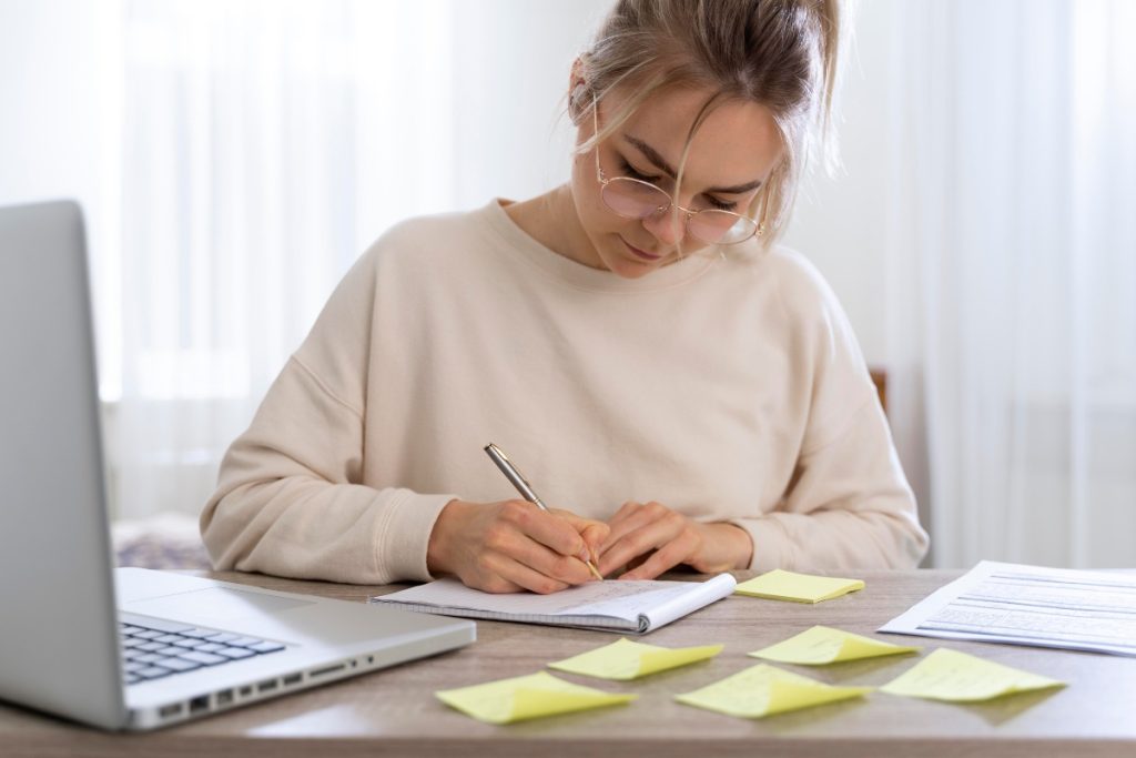 Na imagem, uma mulher está sentada em uma mesa de madeira, concentrada em escrever em um caderno com um lápis. Ela usa óculos e veste uma blusa clara. Na mesa, há vários post-its amarelos espalhados com anotações, um laptop aberto e uma folha com gráficos. O ambiente é claro e iluminado, com cortinas brancas ao fundo. A mulher parece estar em um ambiente de trabalho ou estudo, focada em suas tarefas. Essa é a foto de capa do texto sobre Dicas para Redação.
