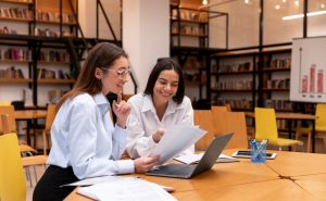 A imagem ilustra duas mulheres praticando a educação continuada, que é o hábito de se manter sempre estudando e se atualizando sobre determinado assunto ou segmento. Elas estão sentadas em uma mesa redonda de madeira clara, ambos estão com camisas sociais lisas claras e estão olhando para papéis e um computador.