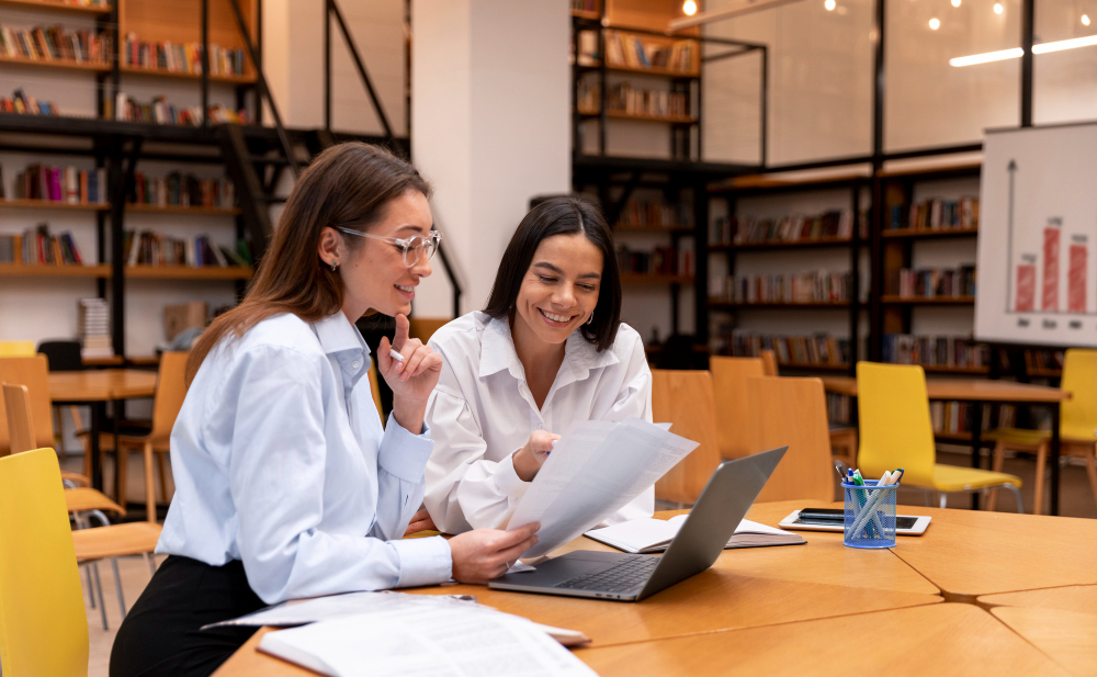 A imagem ilustra duas mulheres praticando a educação continuada, que é o hábito de se manter sempre estudando e se atualizando sobre determinado assunto ou segmento. Elas estão sentadas em uma mesa redonda de madeira clara, ambos estão com camisas sociais lisas claras e estão olhando para papéis e um computador.