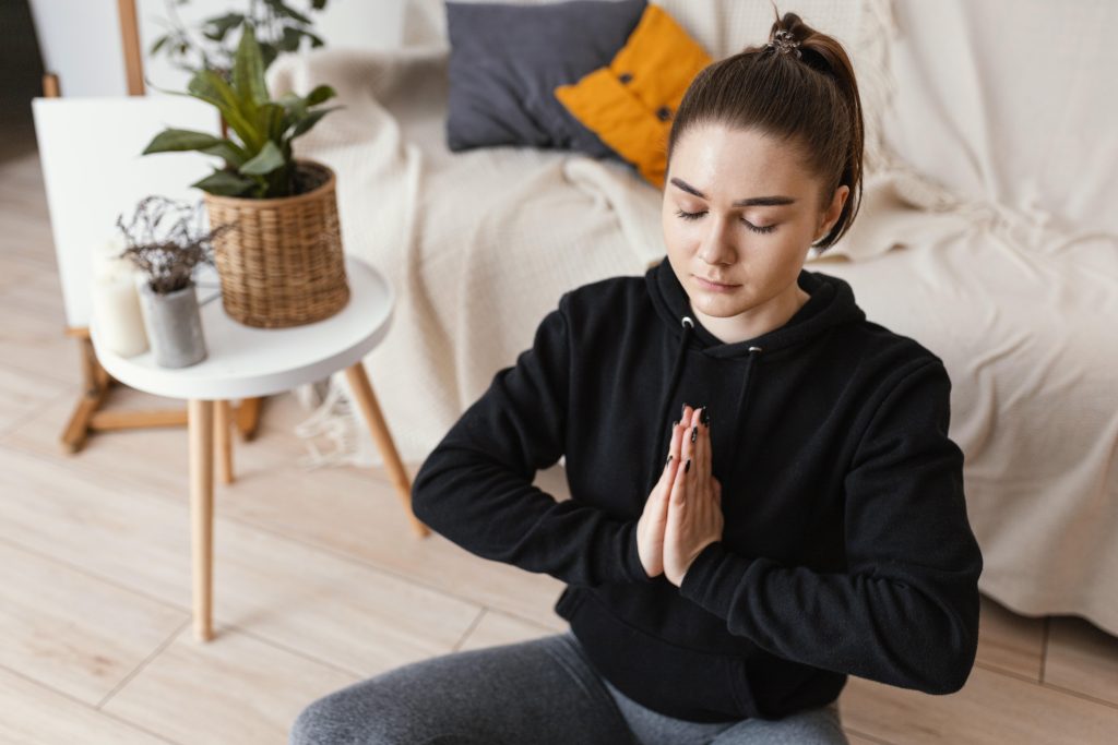 Mulher jovem em posição de meditação com as mãos unidas em prece, praticando mindfulness em um ambiente aconchegante.