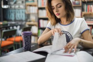 Jovem estudando em uma biblioteca, folheando um caderno enquanto está cercada por livros e com um copo reutilizável ao lado.