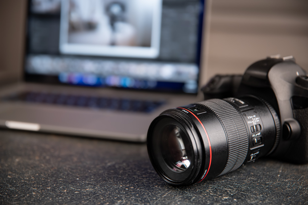 Câmera DSLR com lente profissional em foco sobre uma superfície escura, com um laptop ao fundo exibindo uma tela de edição de imagens.