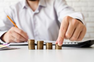 Pessoa com camisa social organizando pilhas de moedas sobre uma mesa, com calculadora e documentos ao fundo, representando planejamento financeiro.
