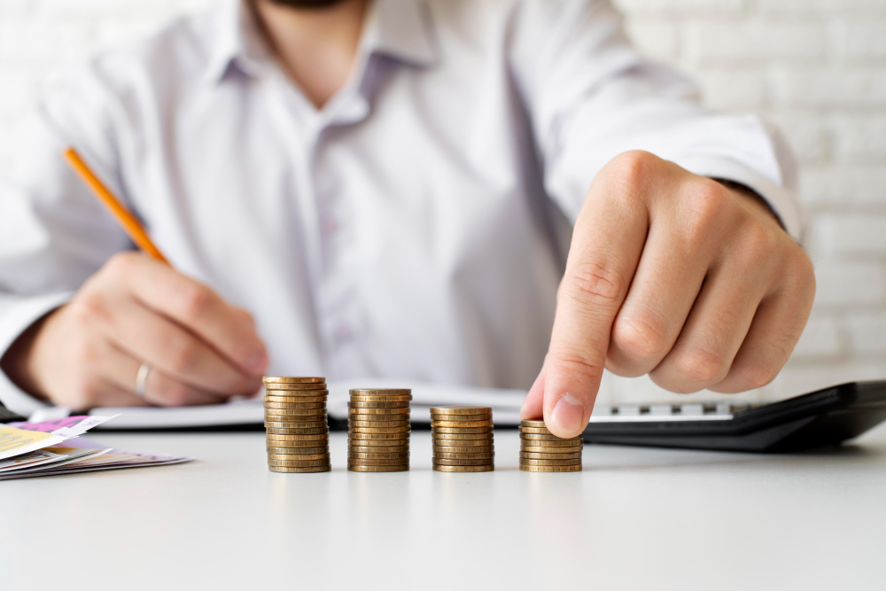 Pessoa com camisa social organizando pilhas de moedas sobre uma mesa, com calculadora e documentos ao fundo, representando planejamento financeiro.
