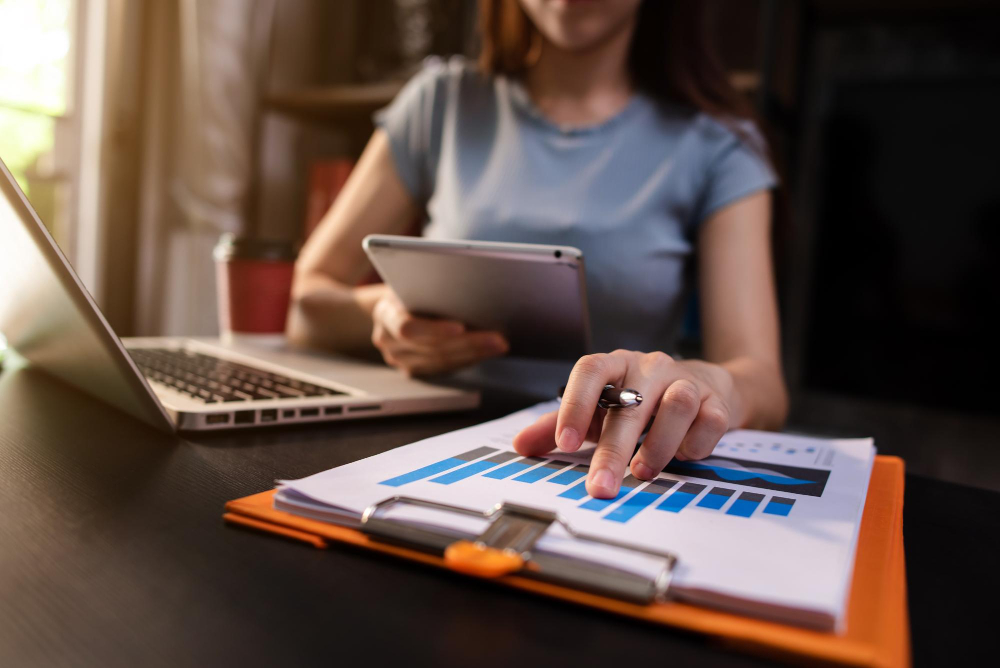 Mulher em ambiente de trabalho analisando um relatório com gráficos de barras, segurando um tablet, com notebook e prancheta sobre a mesa.