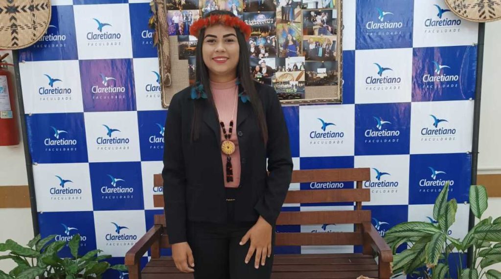 Jovem com vestimenta social e adornos indígenas posando sorridente em frente ao painel com o logotipo da Claretiano Faculdade, com decoração temática ao fundo.