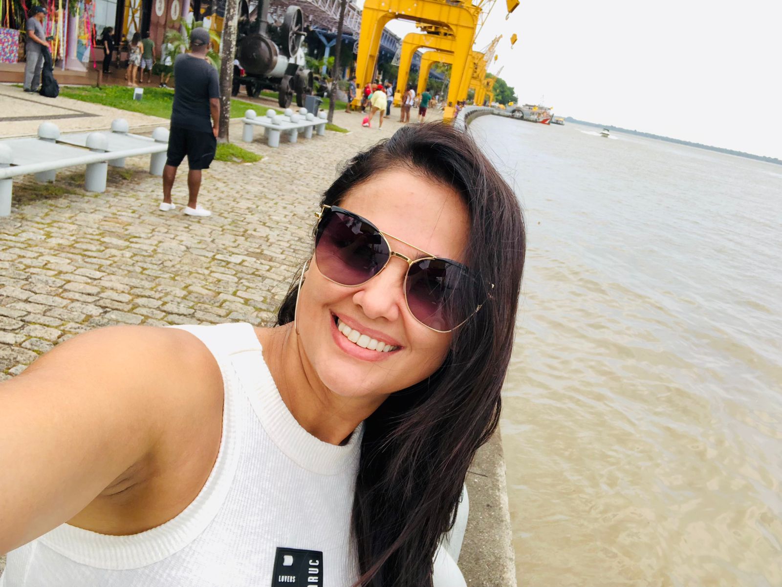 Mulher com blusa branca e óculos de sol tirando selfie em área de passeio próxima ao rio, com guindastes amarelos e pessoas ao fundo.
