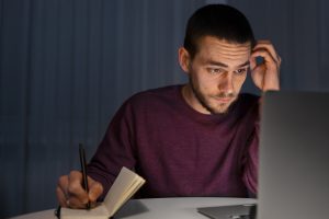 Homem jovem sentado em uma mesa, olhando concentrado para o laptop enquanto escreve em um bloco de anotações à noite.