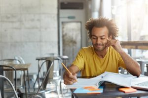 Homem jovem com cabelo crespo e camiseta amarela estudando em um café, com livros, post-its e um laptop sobre a mesa.