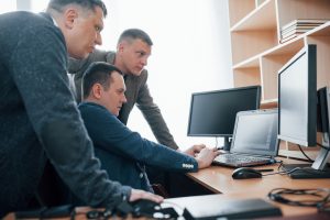 Três homens em um escritório analisam atentamente a tela de um laptop. O homem sentado digita, enquanto os outros dois, de pé, observam a tela com expressão séria. O ambiente tem múltiplas telas, cabos e uma estante com livros.