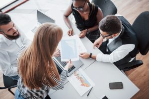 Quatro profissionais em uma reunião de negócios analisando gráficos impressos e trabalhando em laptops ao redor de uma mesa de escritório.