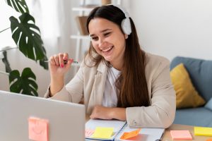 Mulher sorridente com fones de ouvido participando de uma videoconferência, com caderno e post-its coloridos sobre a mesa, em um ambiente iluminado e decorado.