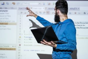 Um homem com barba e moletom azul segura um laptop enquanto aponta para uma tela projetada com linhas de código e interface de um editor de programação. O reflexo do código é visível em seu rosto e roupa.