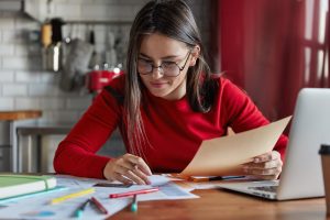 Uma mulher jovem de cabelos castanhos, usando óculos e um suéter vermelho, está sentada em uma mesa de madeira com vários papéis, canetas e um laptop aberto. Ela segura uma folha enquanto analisa documentos com uma expressão concentrada. Ao fundo, há uma parede com azulejos brancos e utensílios de cozinha pendurados.