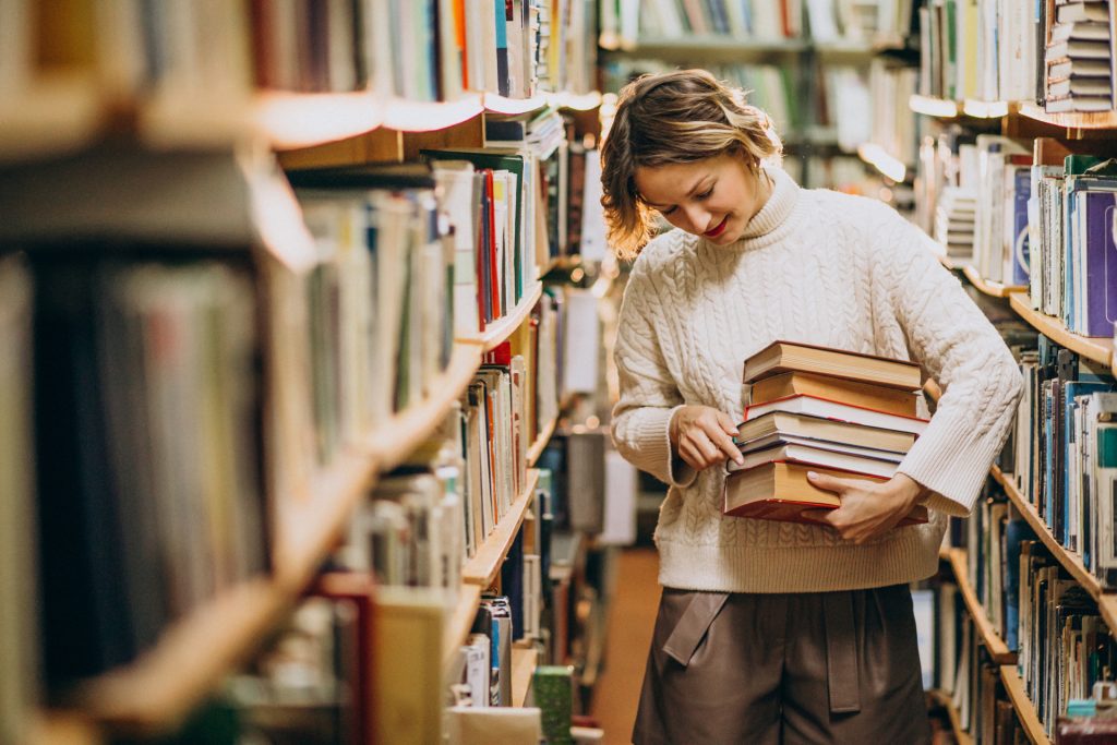 Uma jovem com cabelos curtos ondulados, vestindo um suéter branco de tricô, segura uma pilha de livros enquanto caminha entre prateleiras repletas de obras em uma biblioteca. Ela olha para os livros com um leve sorriso.