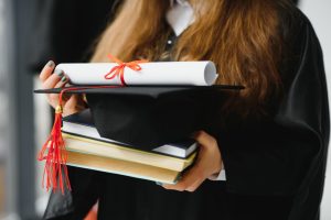 studante em beca de formatura segurando um capelo com borla vermelha, um diploma enrolado e uma pilha de livros, representando a conquista acadêmica.