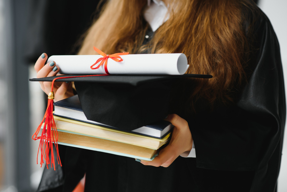 studante em beca de formatura segurando um capelo com borla vermelha, um diploma enrolado e uma pilha de livros, representando a conquista acadêmica.