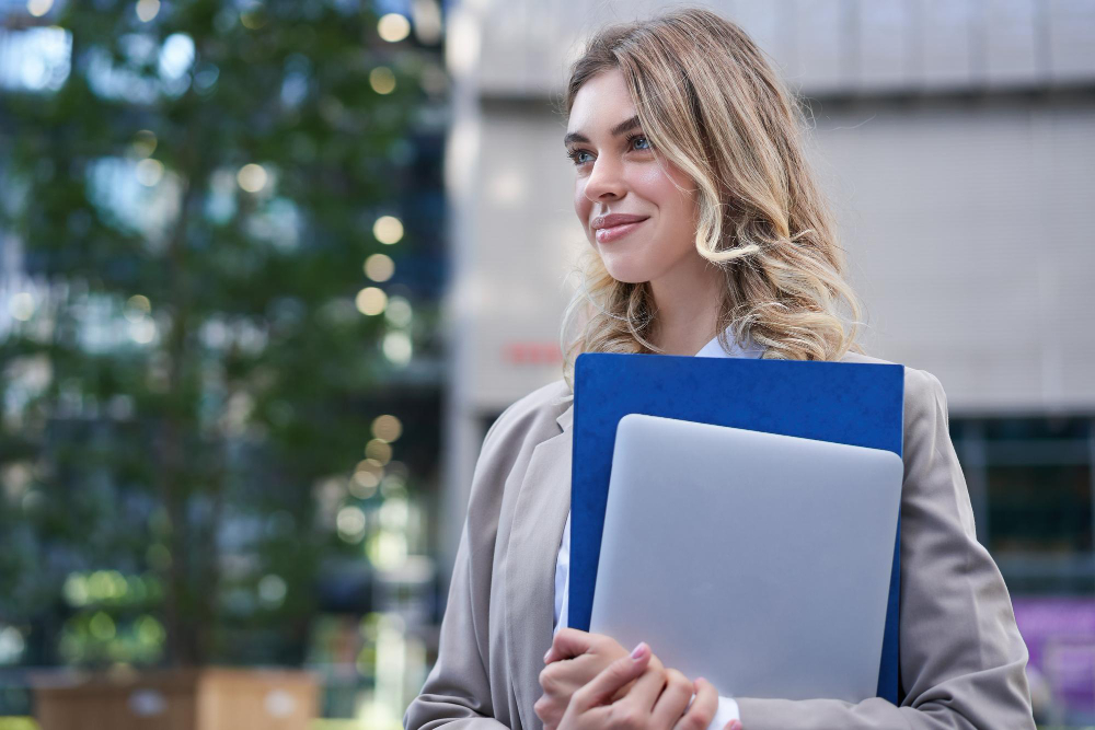 Mulher jovem e sorridente, vestindo blazer bege e camisa branca, segurando um laptop e uma pasta azul em um ambiente urbano com prédio moderno e árvores ao fundo.