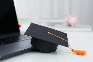 Capelo de formatura preto com tassel laranja apoiado sobre um notebook em uma mesa branca.
