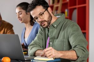 Homem de óculos escreve em caderno enquanto usa notebook, com colegas ao fundo em ambiente de estudo colaborativo.