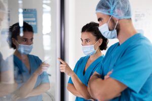 Dois profissionais da saúde, usando máscaras e roupas hospitalares azuis, analisam informações refletidas em uma parede de vidro.