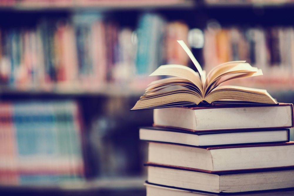 Pilha de livros com um livro aberto no topo, em frente a uma estante desfocada.