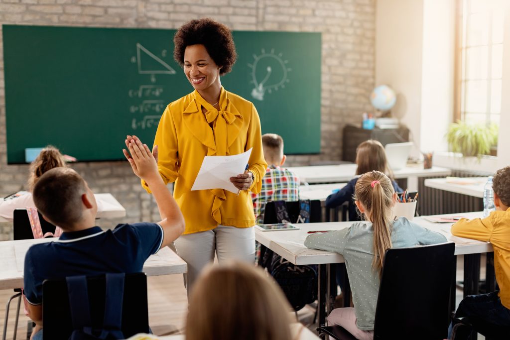 Professora sorridente dá um "high five" para aluno em sala de aula enquanto segura uma prova.