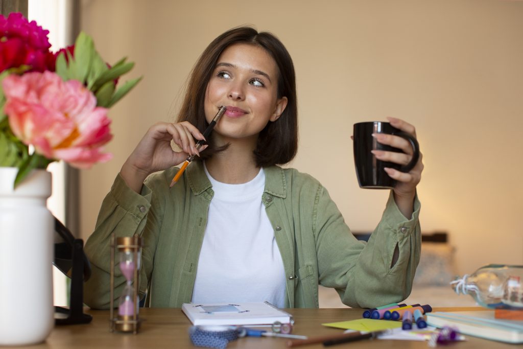 Mulher sorridente segurando caneca e caneta enquanto faz anotações em uma mesa.