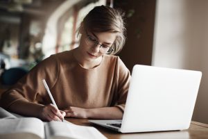 Menina com cabelos louros acobreados e óculos estudando para o vestibular de medicina, com notebook, livro, caderno e lápis sobre a mesa.