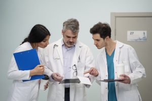 Três profissionais da saúde de jaleco branco analisando documentos juntos em ambiente hospitalar.