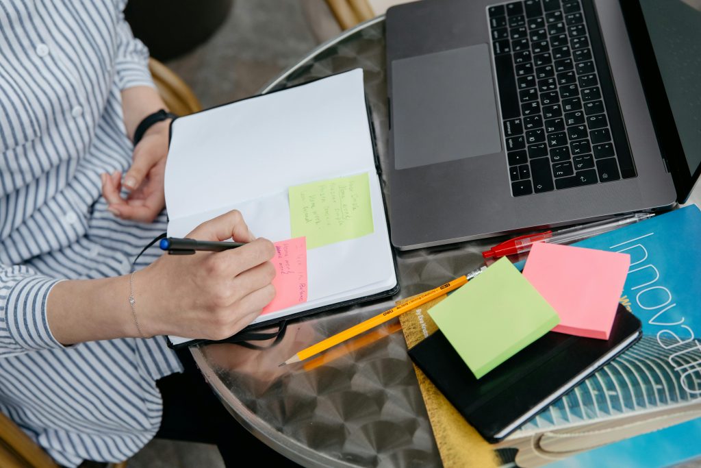 Pessoa fazendo anotações em post-its ao lado de laptop e materiais de leitura, simbolizando estudos de pós-graduação.