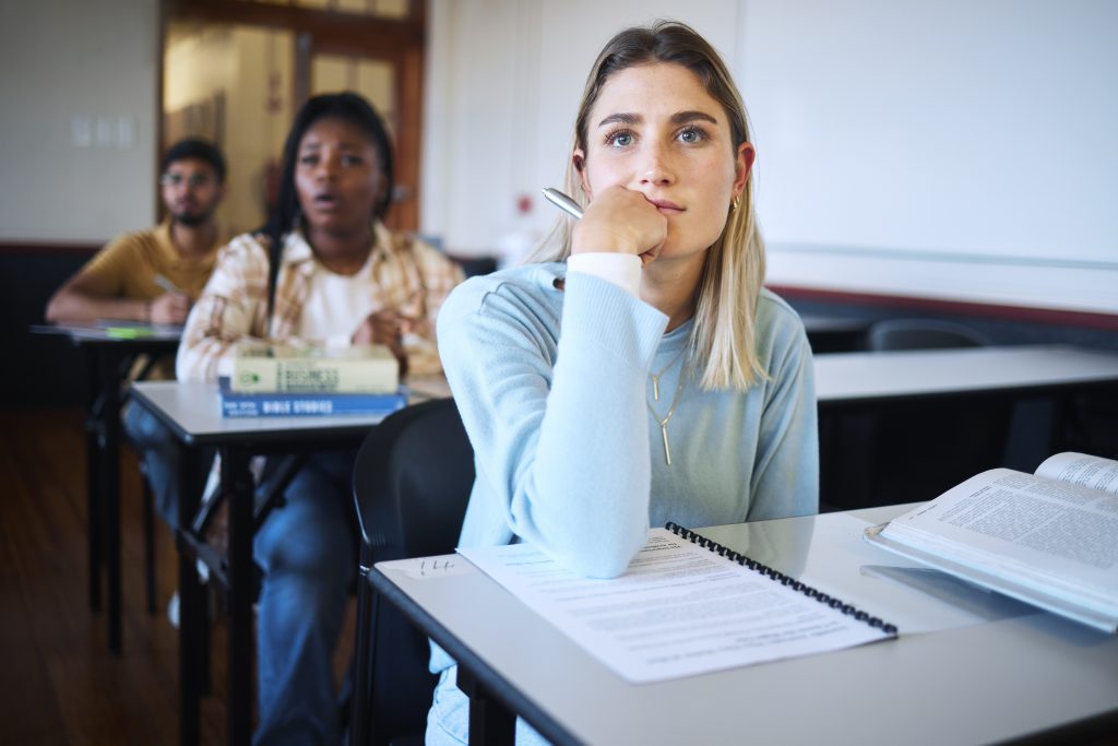 Estudante em sala de aula, pensativa, com materiais de estudo sobre a mesa.