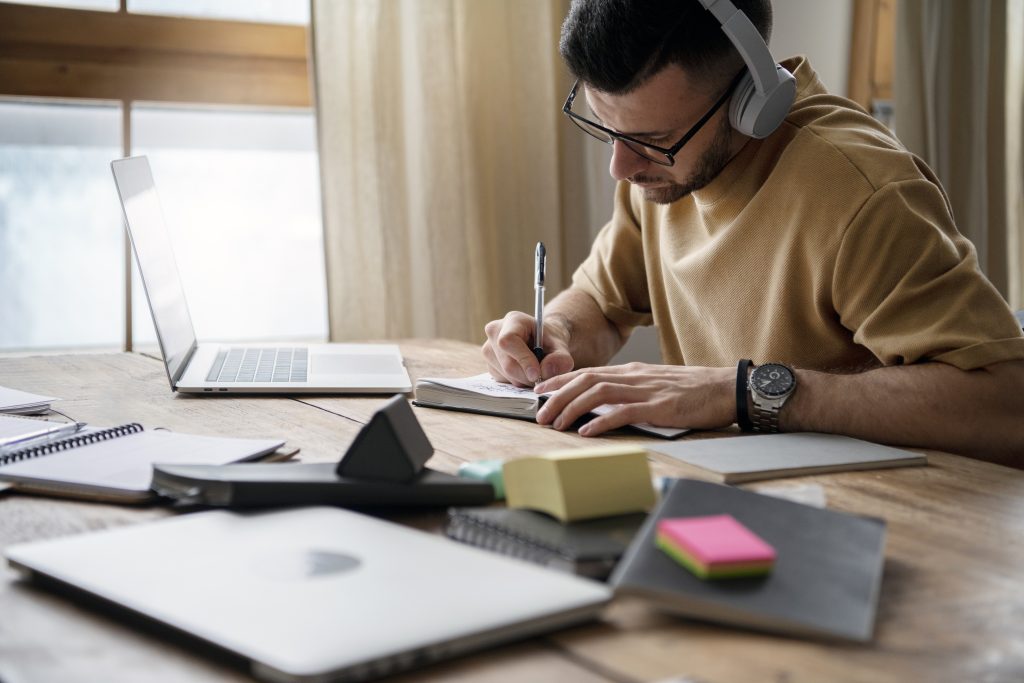 Homem estudando para o vestibular com fones de ouvido, escrevendo em um caderno ao lado de um notebook em uma mesa de madeira.