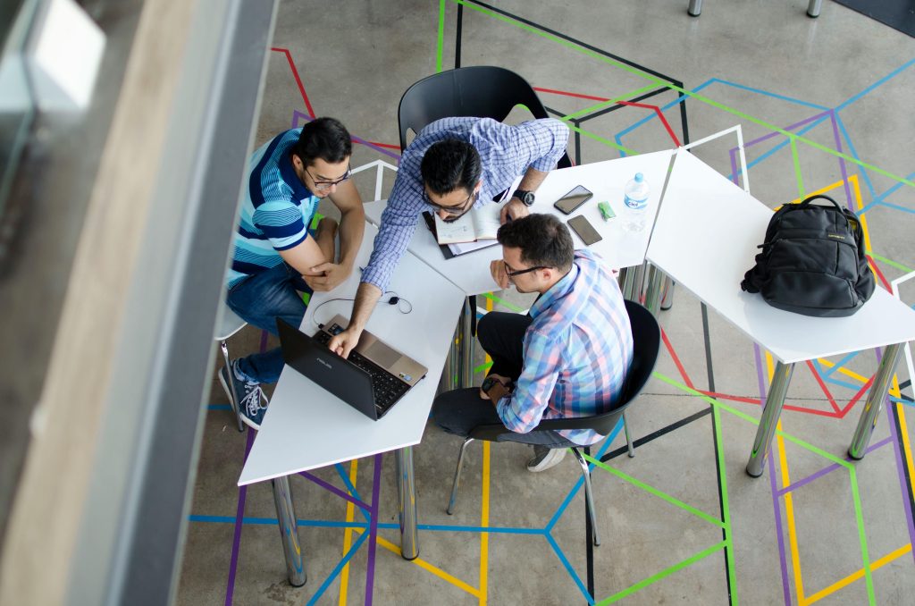 Três homens sentados ao redor de uma mesa em um ambiente moderno, colaborando em um notebook, representando um momento de networking e troca de ideias.